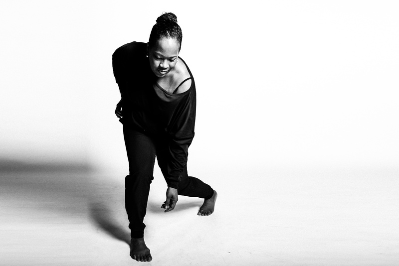  Black woman in black and white photo, wearing a dark leotard, bent forward toward the camera with arm gently stretched down to the floor 