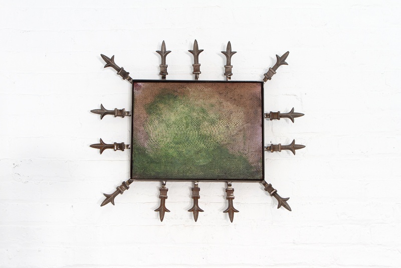  Fabric mounted on the wall on a small black frame. The fabric is green and faded with fence spears surrounding it. 