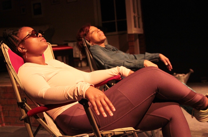  Two performers lounge in chairs on stage. One is wearing sunglasses. 