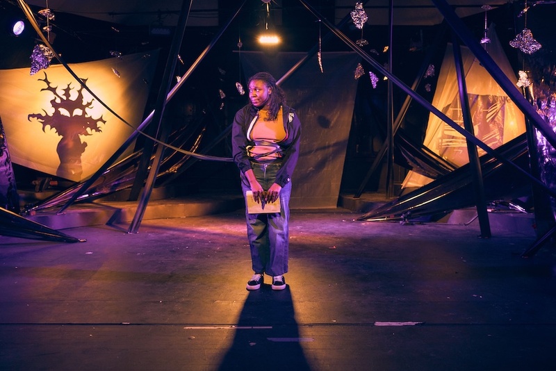  A performer stands on stage holding a piece of paper. They are surrounded by black fabric. 