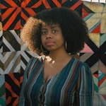 young woman with curly hair and brown skin stands in front of a colorful tapestry