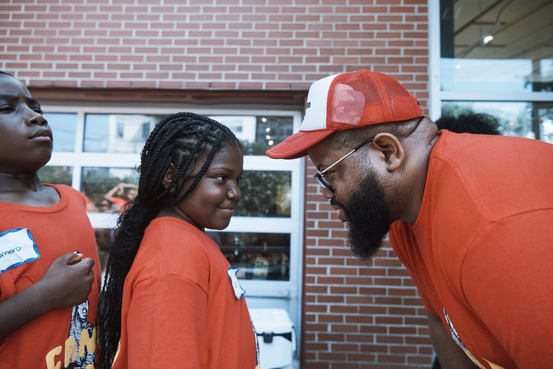  A Black man with a red hat on and red shirt leans forward talking to a small girl who is smiling. 