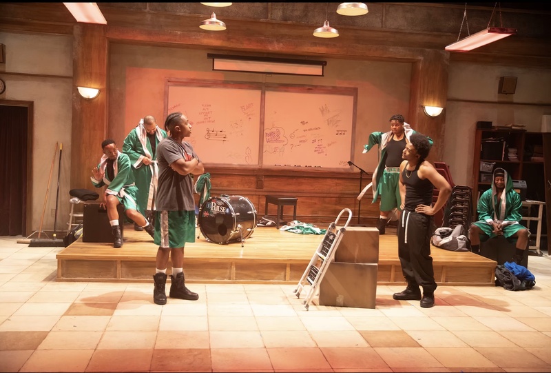  Young black men in green and black athletic leisure floor stand in a band room. There are mismatched tan tiles on the floor, an old wooden stage, and a whiteboard with lots of drawings behind them. Several hanging gray pendant lights hang overhead. 
