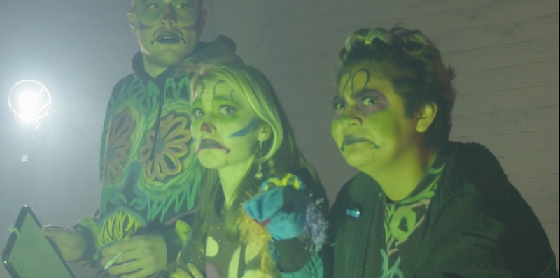  Three performers with clown makeup make funny faces at the camera. One performer has a hand puppet. 