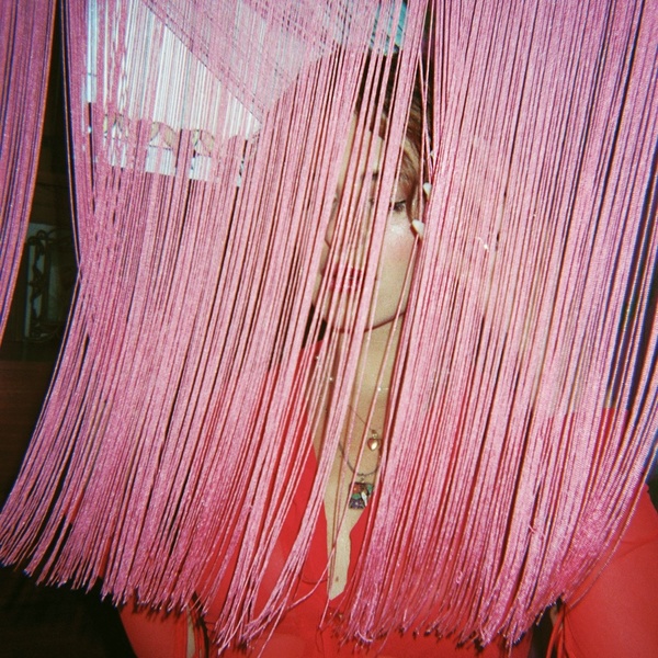  A femme-presenting person with long curly dark hair poses behind a frilly pink curtain 