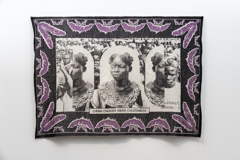  A photograph of a textile hung on a white wall. The textile is black with purple feathered detailing surrounding the edge. In the center a Black woman is woven three times with the name CHEMA CHAJIUZA KOBAYA CHAJITEMBEZA. 