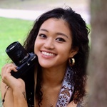 asian person with dark brown hair and brown eyes with camera leaning on her chin smiles largely at the camera
