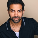 Headshot of Richard Costes, a brown man with black hair, stubble flecked with gray, and brown eyes. He is wearing a black polo shirt slightly unbuttoned to show a necklace and a white shirt underneath.