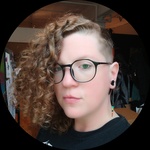 A white, queer androgynous person with a red curly hair, a close cropped side, glasses and grey eyes stands in front of their studio.