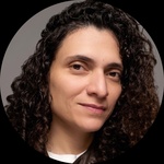 A Latin American person with long, dark, curly hair and brown eyes sits in front of a neutral background wearing a brown leather jacket; she uses she/they pronouns.