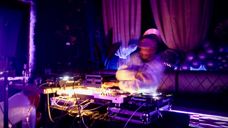  A distorted photo of a DJ playing in a purple lit room. The DJ is moving so their frame is blurred, they are standing in front of a folding table that has an array of DJ equipment. 