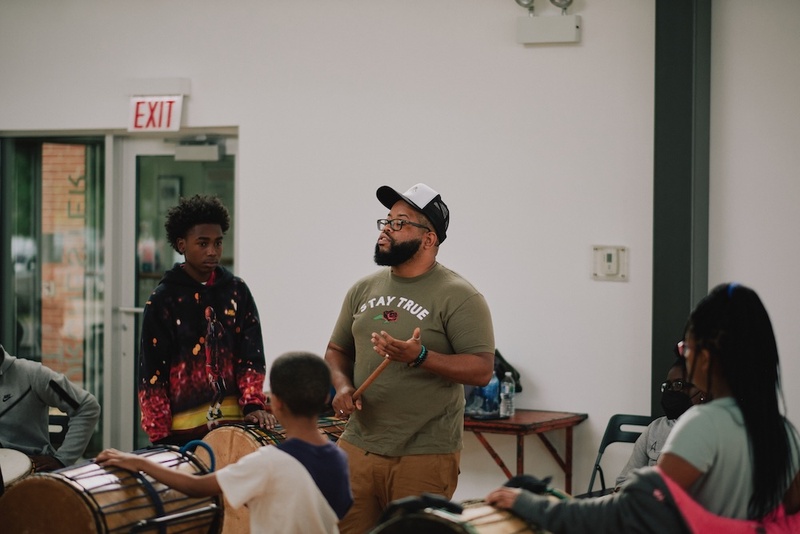  A Black man holds a drumstick while instructing a group of kids how to play the drums/ 