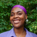 Image of Simone smiling, nestled in a tree. She is wearing a purple head wrap, lavender button up, silver hoops, and a silver necklace.