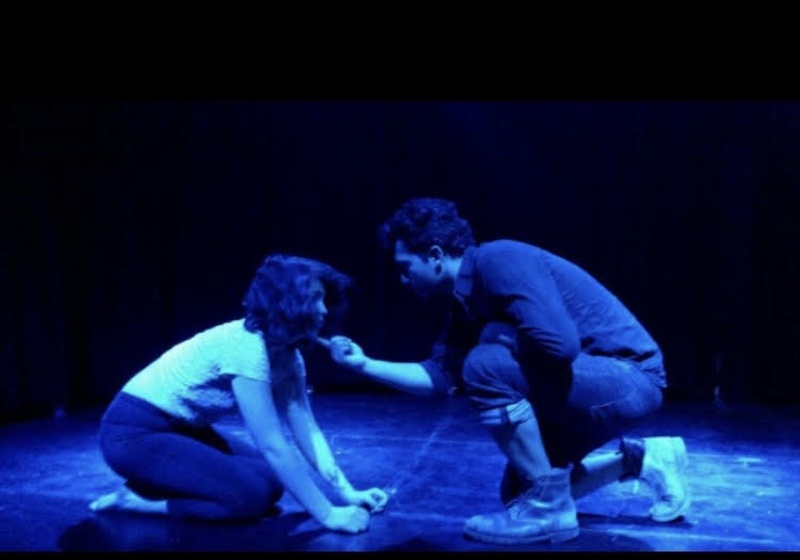  A man and woman are crouched on a stage bathed in blue light 