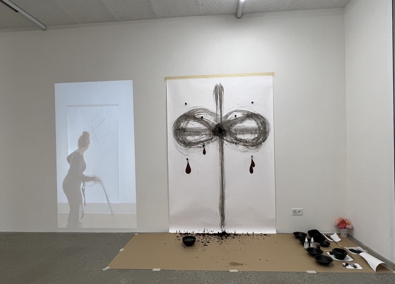  Gallery view showing a looped 17 minute video of Rope Beating Drawing projected life-size onto the left and a large white sheet of paper on the right side with a drawing resembling a dragonfly and figure "8" motion made with herbs, soil, oils, and water 