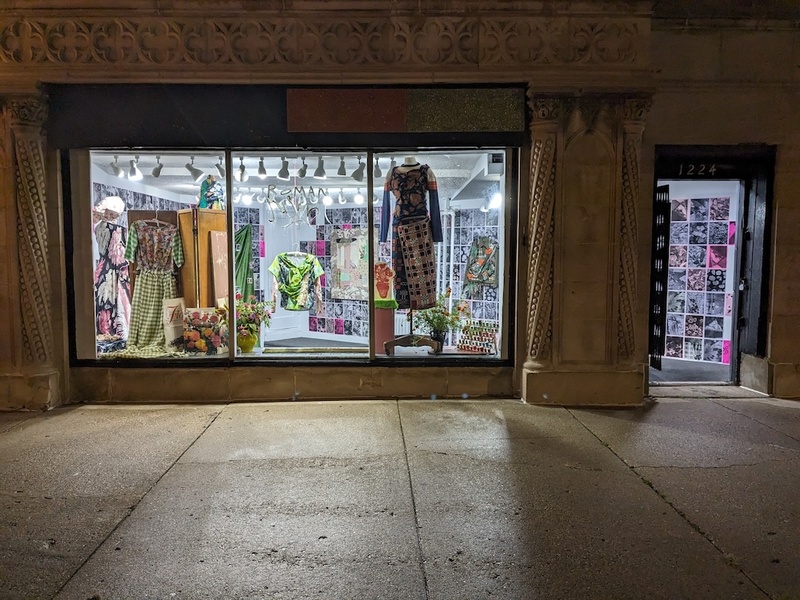  An image two dresses and a shirt hanging in a window. The image is taken from the street, peaking into the gallery. The dresses are made with mismatched fabric in green, blue and patterned materials. 