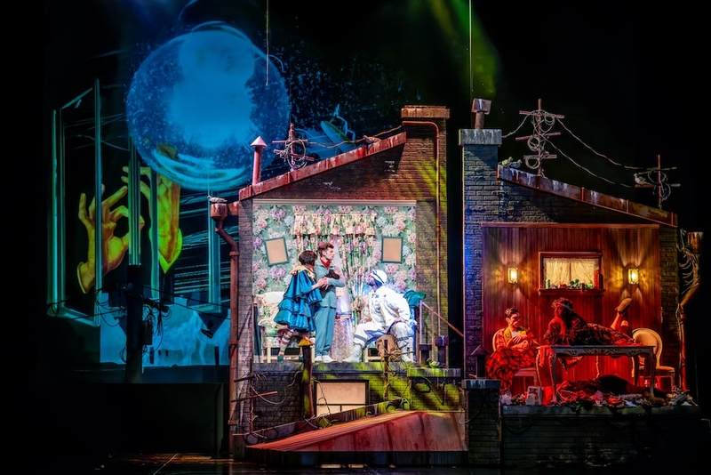  The set of a house split in two, one is indoors and the other is outdoors. On top of the house and in the backdrop there are projections of hands, the sky and the moon. 