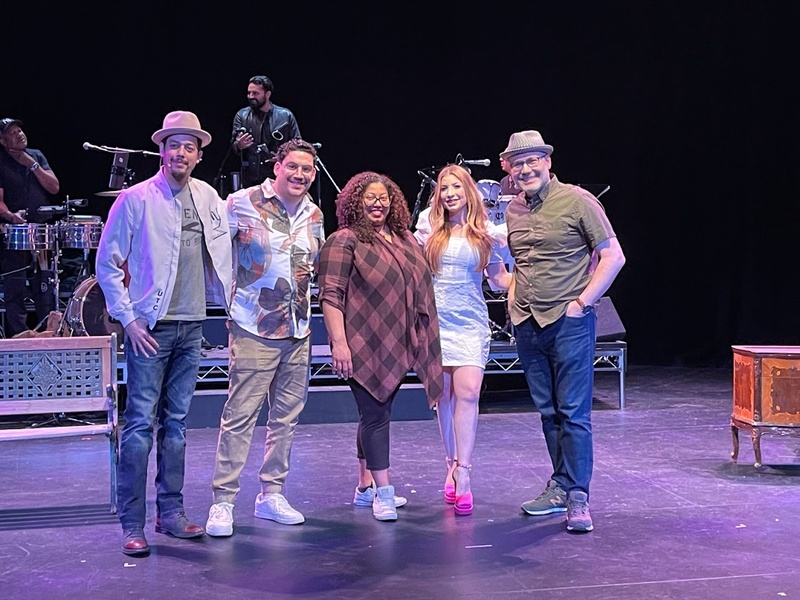  Five people stand on a stage lit in purple light. They are all smiling at the camera with musicians set up in the background. 