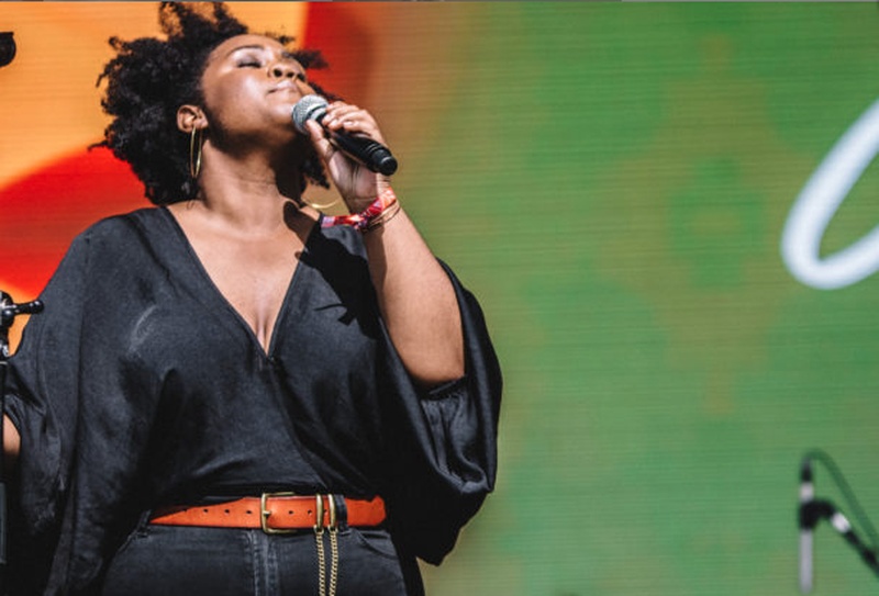  Black woman with curly hair sings into a microphone, her eyes closed and face lifted up 