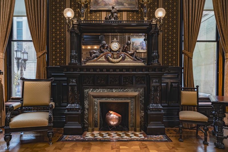  A large, hammered copper bowl filled with copper-gilded coal sits inside a dark, ornate fireplace. Light shines through the inside, illuminating the gilded coal and leaking out of many small holes in the sides of the bowl like constellations. 