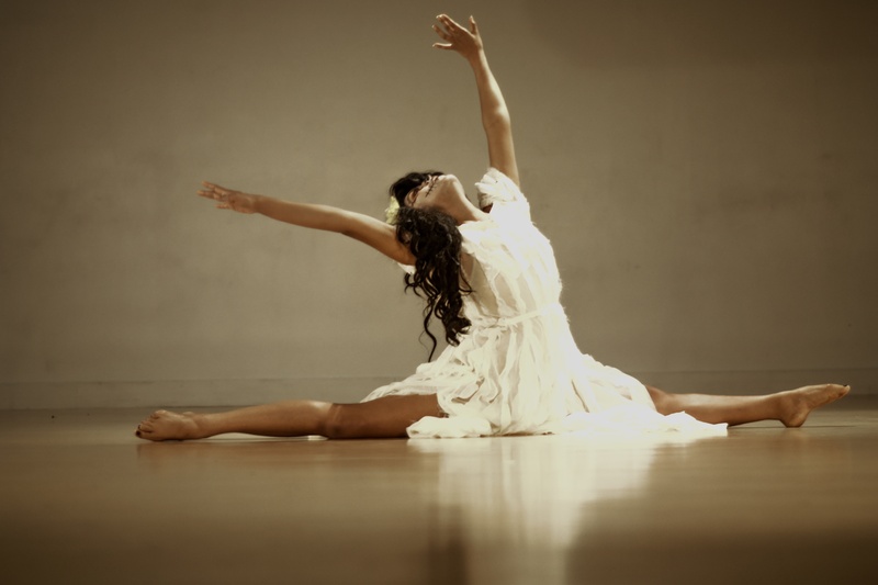  A slender, brown-skinned woman wearing a white dress poses in a split with their arms raised. 