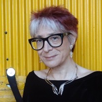 A midddle-aged white woman with red-and-silver streaked hair, black glasses, black sweater, against a yellow door.