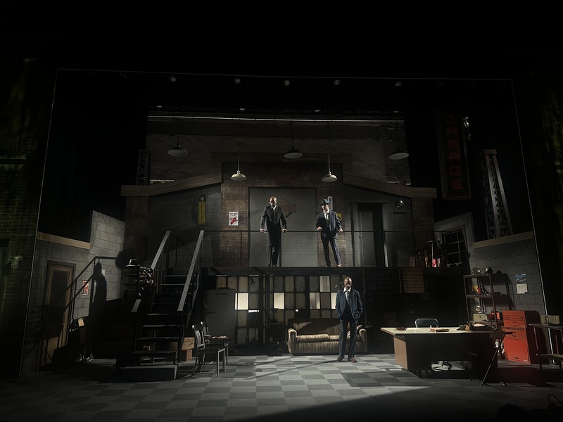  A dark photo of a mechanic's garage with checkered floor. 2 men in suits stand on a second level with pendant lights overhead and a metal staircase to their left. The ground floor has panelled dirty windows, a sofa, a desk, and a man in a suit standing. 