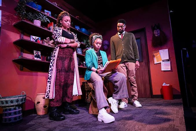 Three performers post on stage. One is seated and reading off an orange folder, the other two look over their shoulder. 