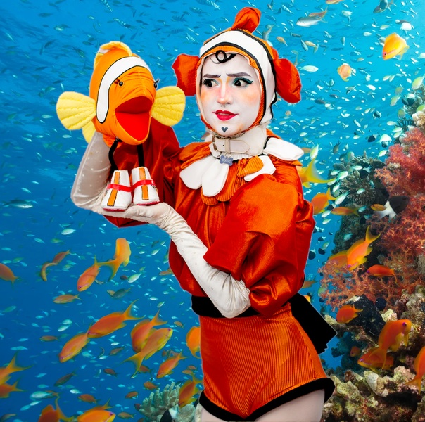  A white, queer transmasculine person dressed in drag as an orange clownfish has a clownfish puppet and is posed in front of a coral reef backdrop. 