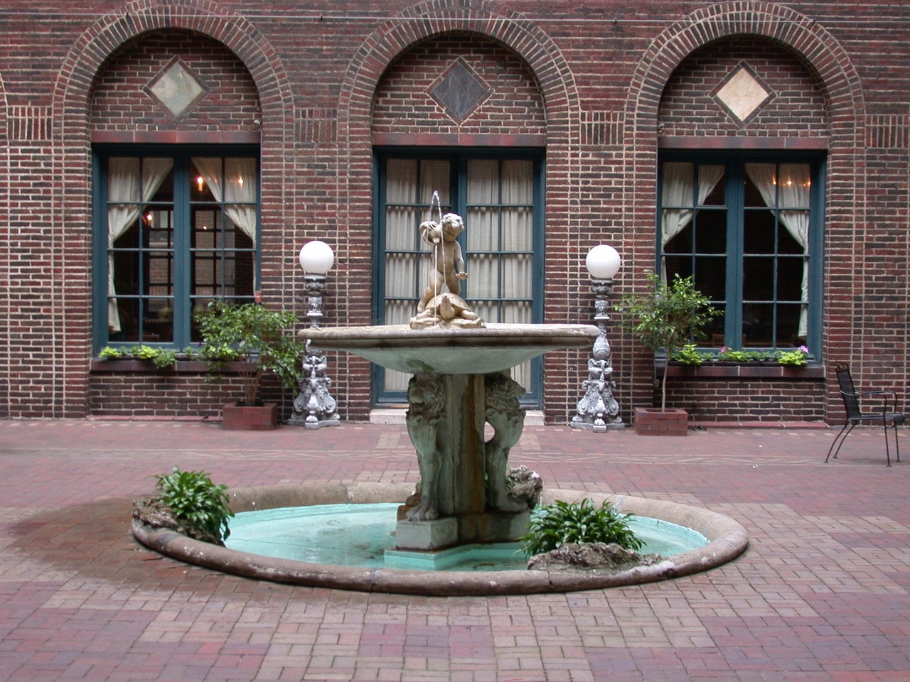 Decorative fountain in the center of a brick patio with three arched windows in the background
