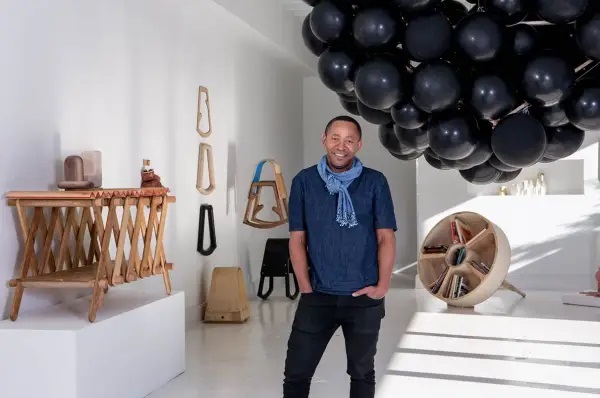 Image of Norman in his exhibition. Furniture objects surround him. He smiles with his hands in his pockets.