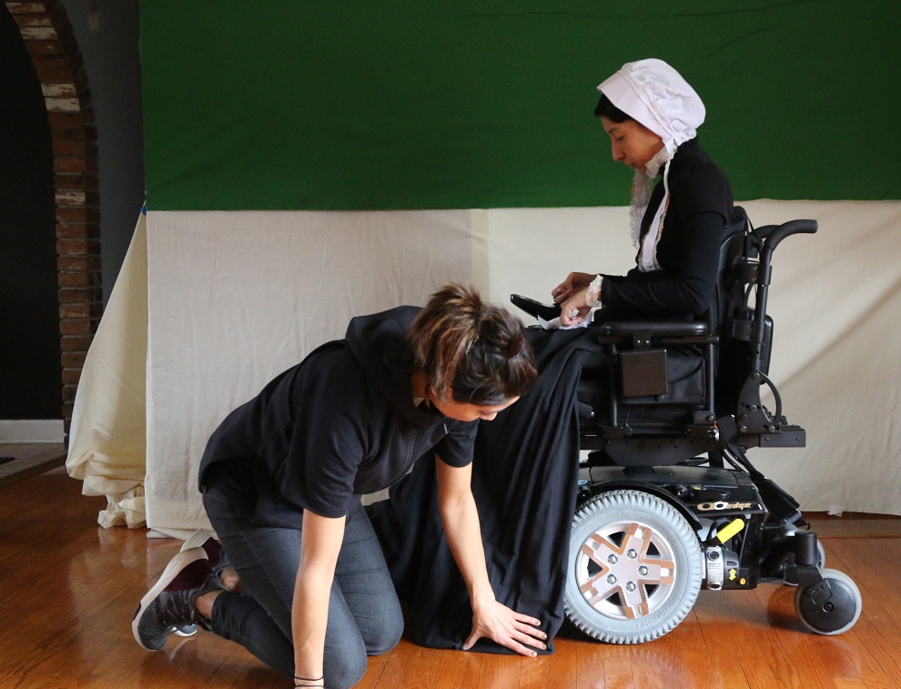 A behind the scenes shot of Reveca Torres (2018 Fellow), a brown skinned woman seated in a wheelchair. Reveca wears a long, black dress and white bonnet for the Tres Fridas Project’s take on Whistler’s Mother (1871) by James McNeill Whistler. An assistant