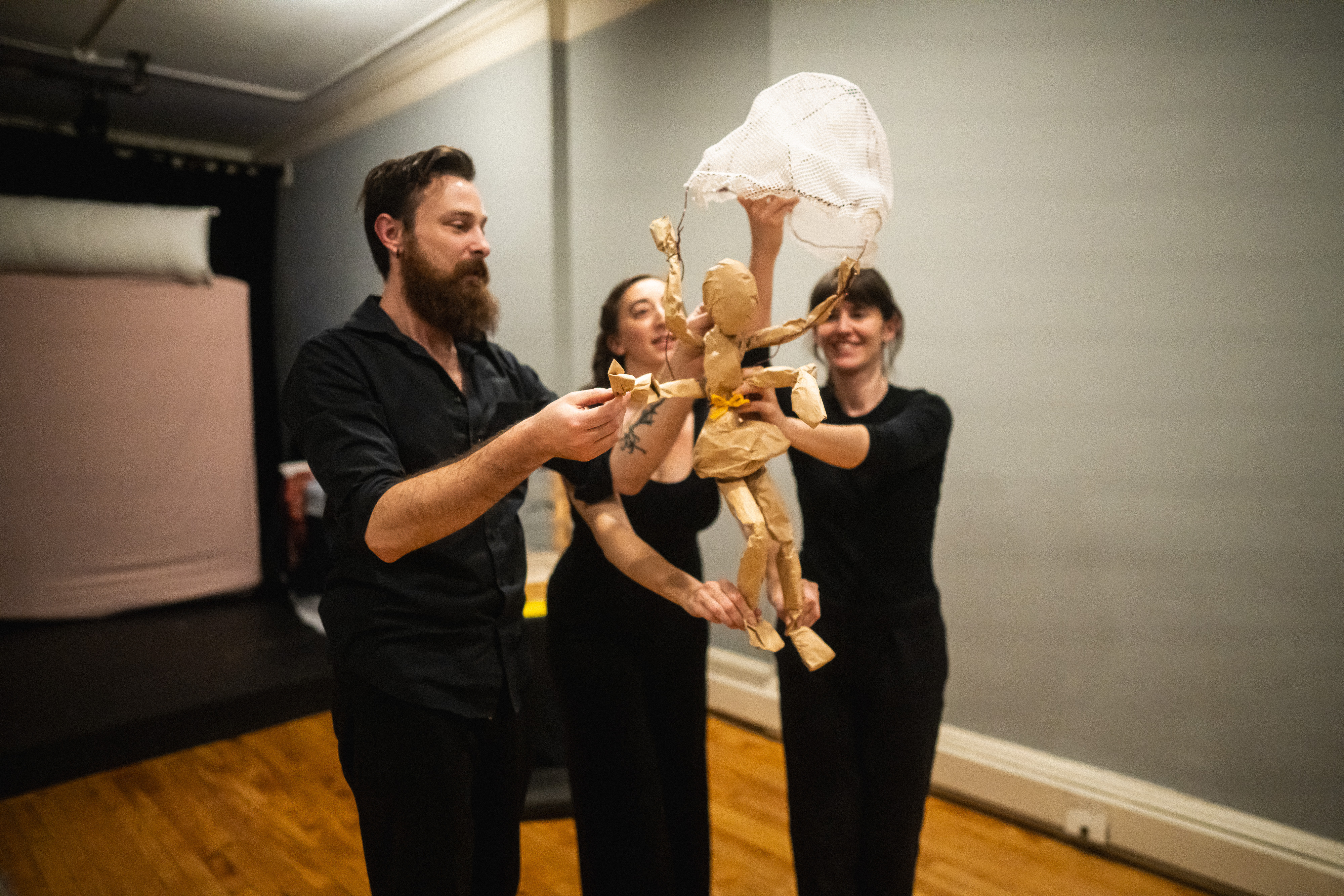 Rachel Singer and Richie Schiraldi demonstrate a tabletop puppetry technique, animating a simple brown paper puppet standing on a table.  The brown paper puppet, around 1.5 to 2 feet tall.  It is a simple form of a body with no defined face.  Rachel holds the back of the puppet head and right arm and Richie holds the feet and left arm to bring the puppet to life.  An ASL interpreter stands to the side of the table.