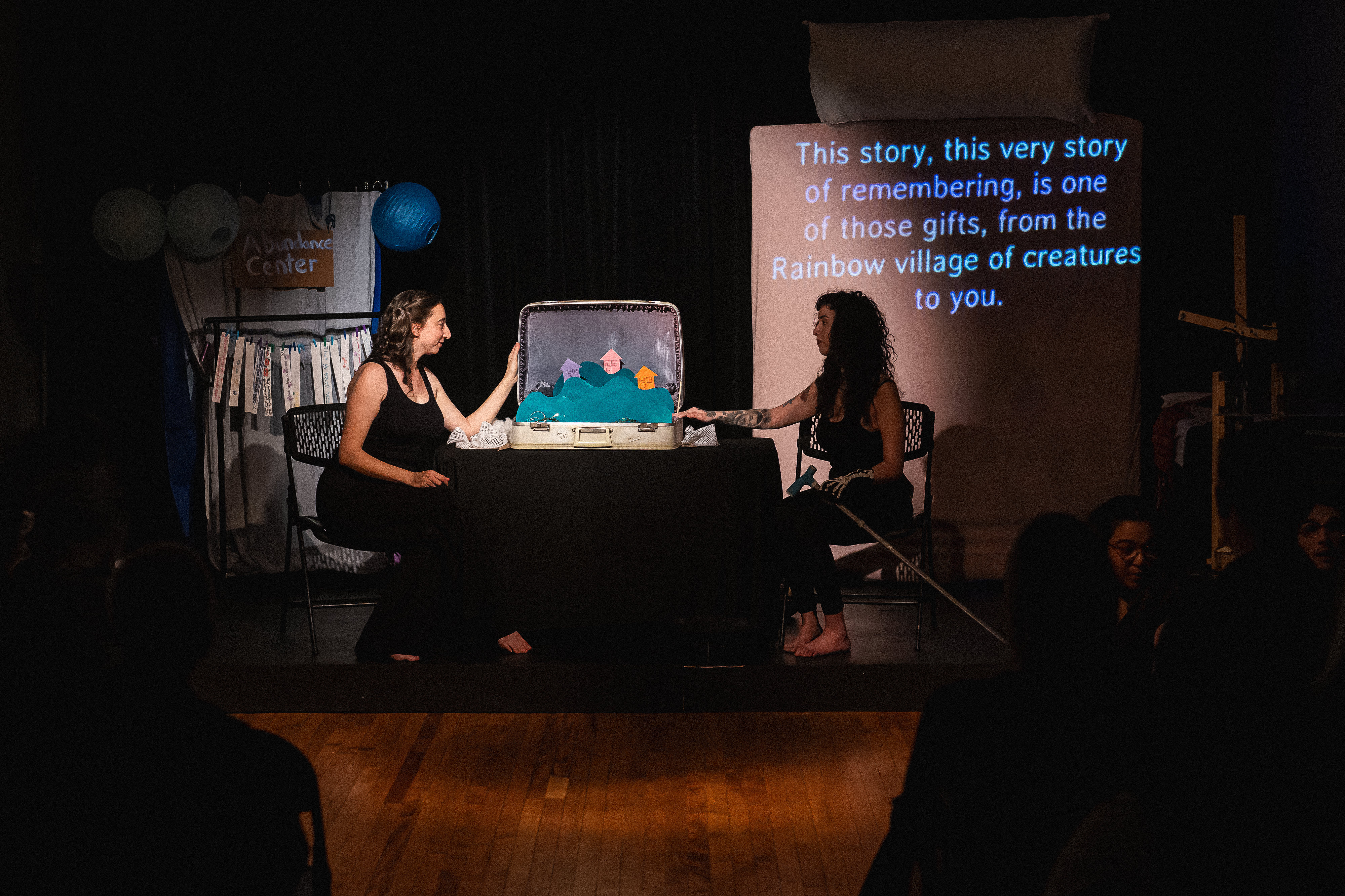 Rachel Singer sits in a chair stage right, Genevieve Ramos sits in a chair stage left.  They look at each other. Between them on a table is a large pop-up book style, two-dimensional colorful village on green paper hills rising out of an open suitcase.  A projection behind Genevieve on the vertical bed has a projection of text that reads: This story, this very story of remembering, is one of those gifts, from the Rainbow village of creatures to you.