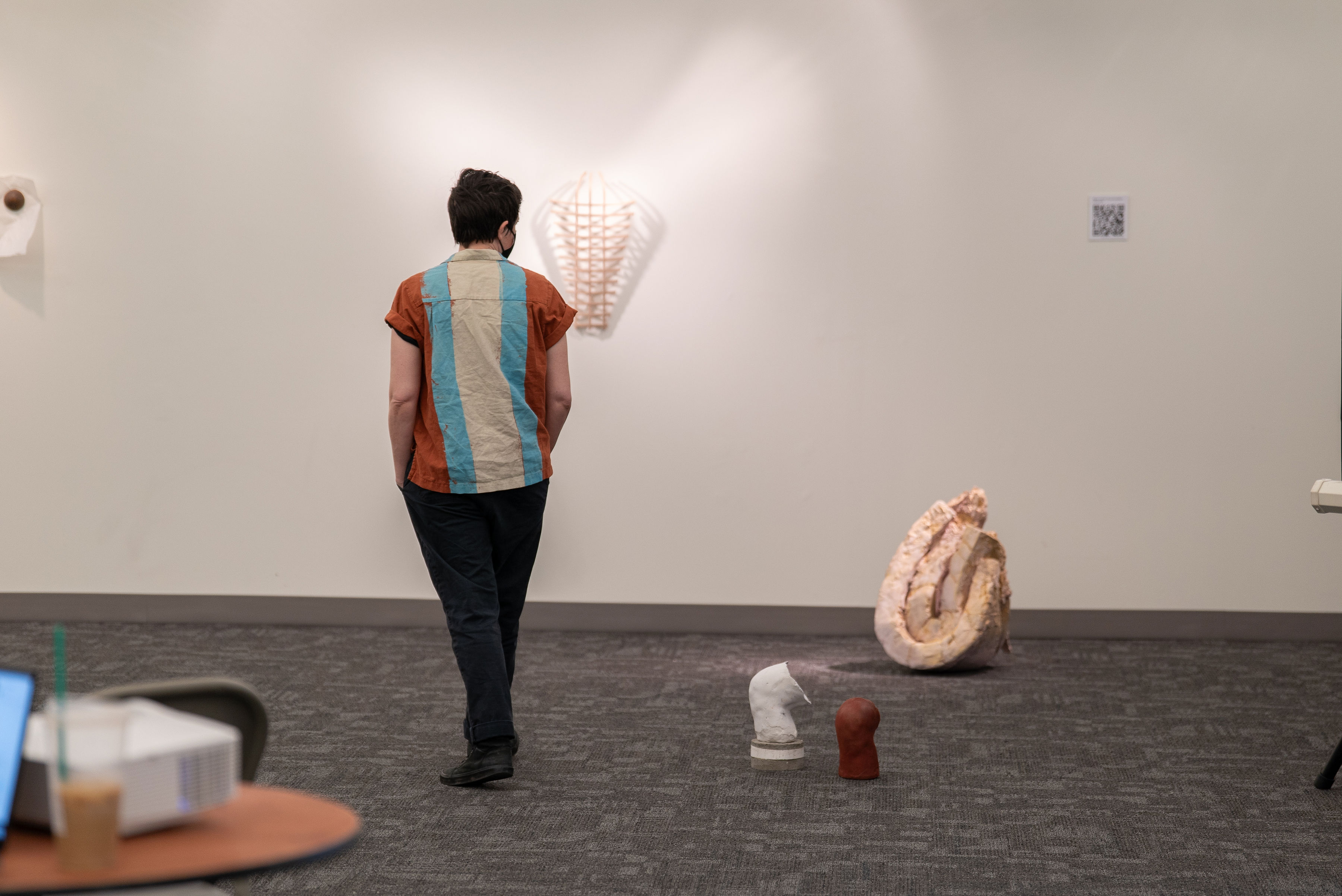 A white person wearing a striped shirt stands in the gallery space with their back to the camera. Blurred in the foreground is a wooden table with a projector on it. The person is standing amongst two sculptures resting on the gray carpeted floor and is gazing at a wooden sculpture hanging on the wall.