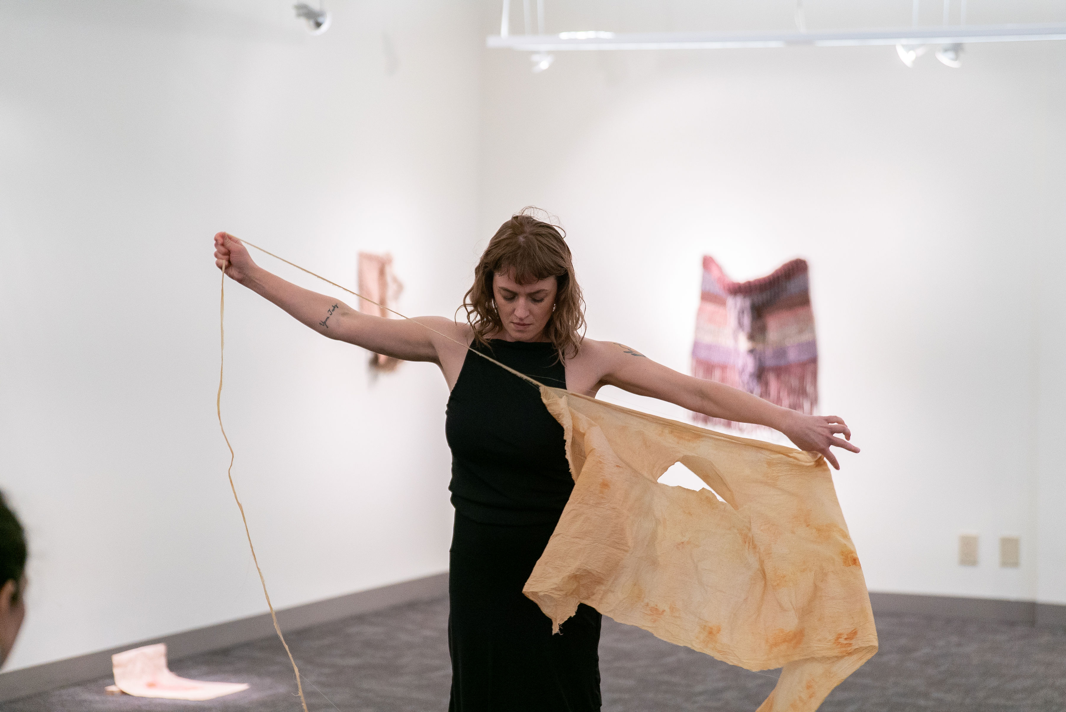 A white woman stands in a white gallery space. Her arms are completely out stretched with one hand gripping a thin strip of fabric and the other end holding a large piece of fabric in a tight tension that allows her to rip the small strip of fabric from the large piece. She is gazing down at the gray carpeted floors.
